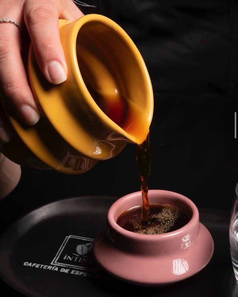 Café de especialidad servido desde jarra cerámica amarilla a taza rosa sobre bandeja Íntimo.