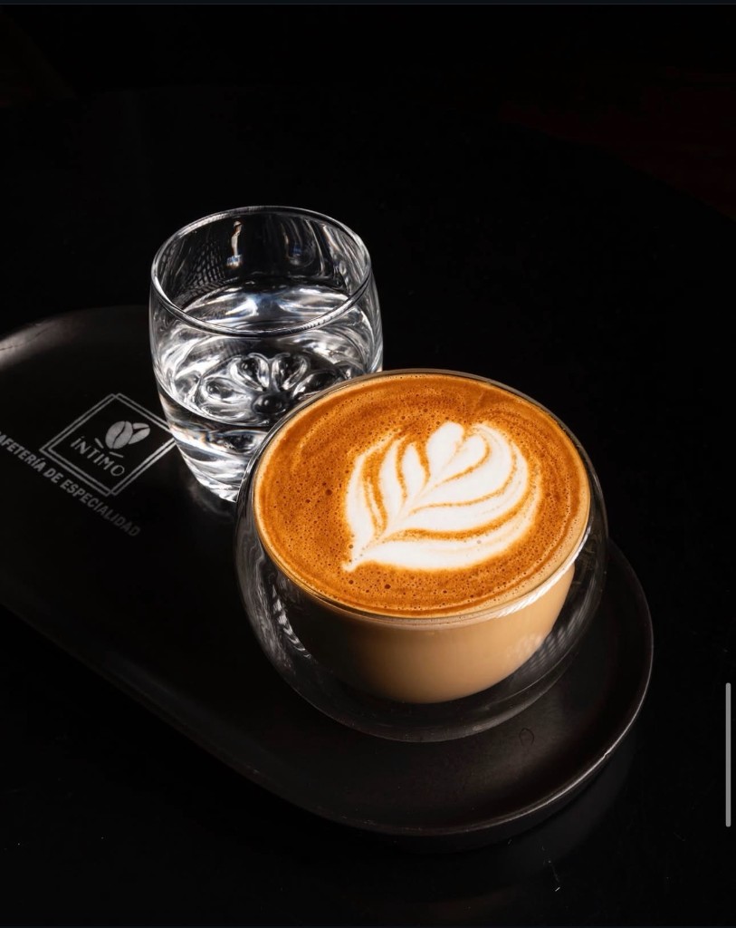 Latte con arte en vaso de doble pared, bandeja negra con logo y vaso de agua.