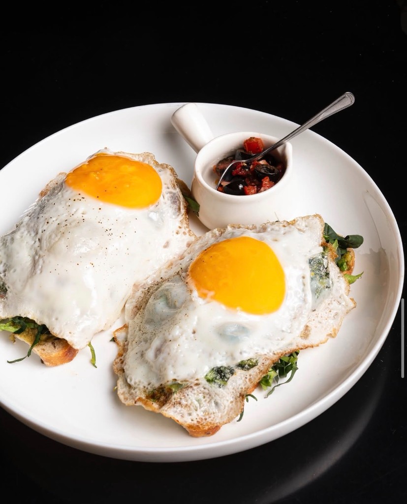 Huevos estrellados sobre tostadas artesanales con verduras y guarnición en plato blanco.